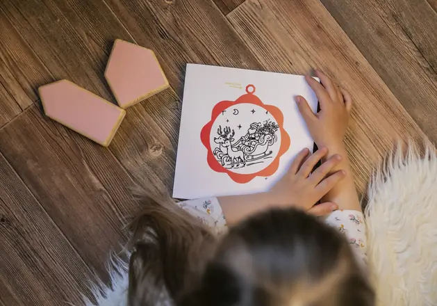 Niña con la página «Para colgar» del libro Navidades Mágicas: un adorno con Papá Noel en su trineo y renos para colorear.