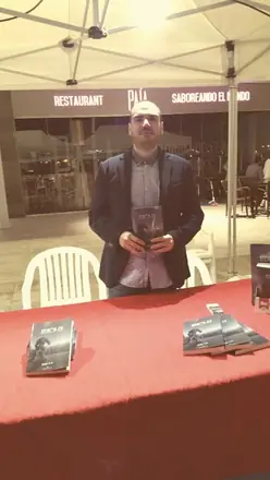 Dan S. V. con un ejemplar de Xerhon-Ra en las manos, de pie tras una mesa de mantel rojo con más libros, al aire libre.