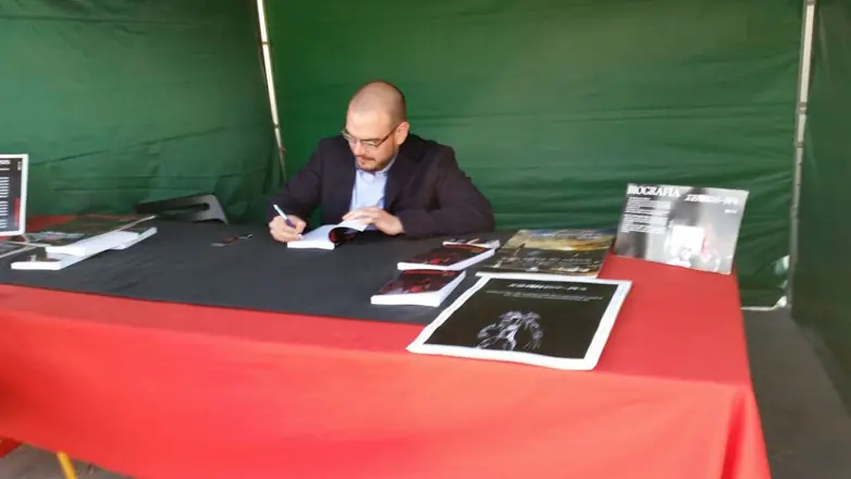 Dan S. V. dedicando un ejemplar de Xerhon-Ra en una caseta verde, con una mesa de mantel rojo y carteles de biografía y sinopsis.