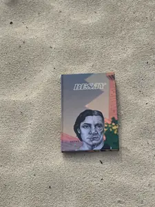 Ejemplar de Besay en tapa dura sobre arena; la portada muestra un rostro dividido entre la juventud y la vejez y el título en letras blancas.