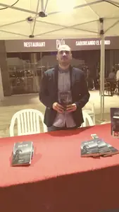 Dan S. V. con un ejemplar de Xerhon-Ra en las manos, de pie tras una mesa de mantel rojo con más libros, al aire libre.