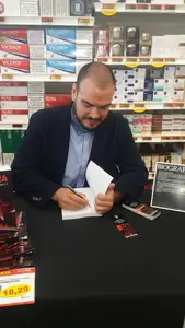 Dan S. V. dedicando un ejemplar de Xerhon-Ra en una mesa con pilas de libros y marcapáginas, en un supermercado.