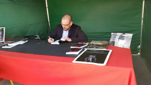 Dan S. V. dedicando un ejemplar de Xerhon-Ra en una caseta verde, con una mesa de mantel rojo y carteles de biografía y sinopsis.