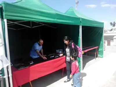 Caseta de firmas con carpas verdes y una mesa de mantel rojo con ejemplares; una mujer con un niño se acerca a la mesa mientras un hombre atiende al fondo.