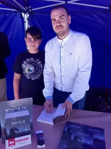 Dan S. V. firmando un ejemplar de Xerhon-Ra bajo una carpa azul, junto a un joven lector; sobre la mesa, un cartel de «libro recomendado» y un marcapáginas.
