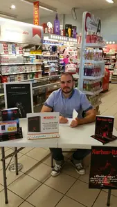 Dan S. V. firmando ejemplares de Xerhon-Ra sentado a una mesa en un supermercado, con el cartel de la firma de libros.