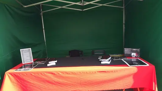 Caseta verde vacía con una mesa de mantel rojo, ejemplares de Xerhon-Ra y carteles de sinopsis y biografía, lista para una firma.